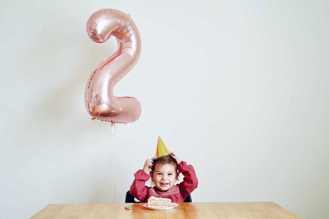 Toddler in a party hat celebrating her second birthday with a 2-shaped balloon and slice of cake.