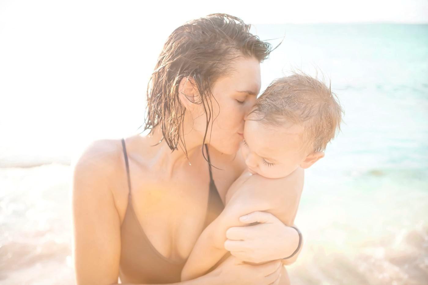 Mother in swimsuit hugging and kissing toddler son on the beach.