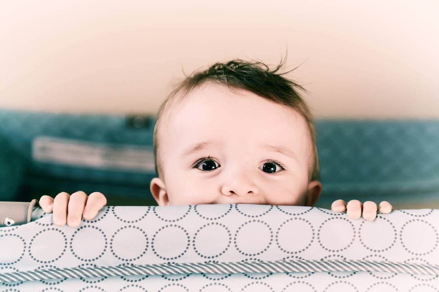 A baby peeks over the side of a portable cot